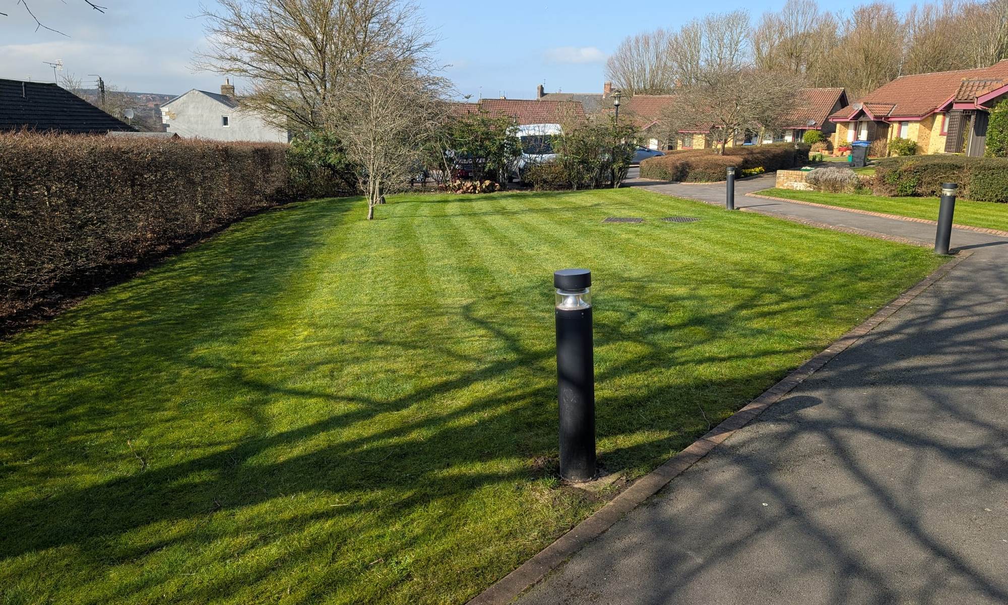 Communal estate lawn mown in stripes beside a tarmac path and a bollard light, bungalows behind