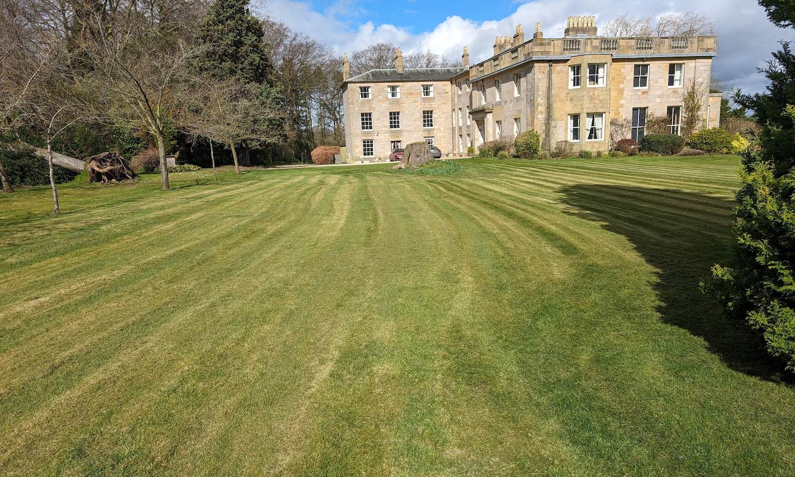 Striped lawn in front of a large stone country house with mature trees
